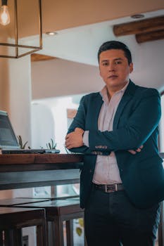 A businessman in a blazer stands confidently in an office with a laptop, embodying professionalism.