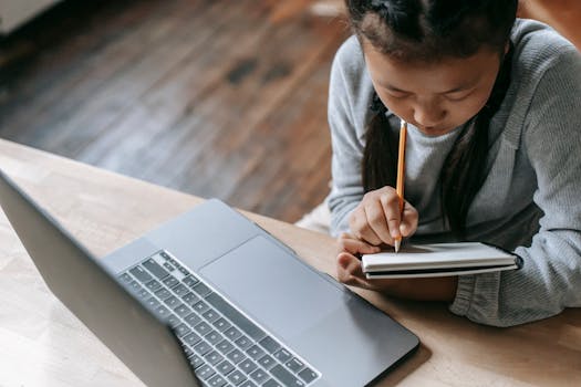 From above of crop attentive ethnic schoolchild writing in workbook with pencil while studying at table with netbook