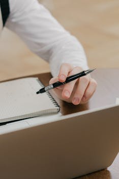 Hand holding a pen over a notepad near a laptop, capturing a work or study moment.