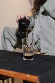 Person in pajamas pouring coffee from a moka pot into a glass in a cozy indoor setting.